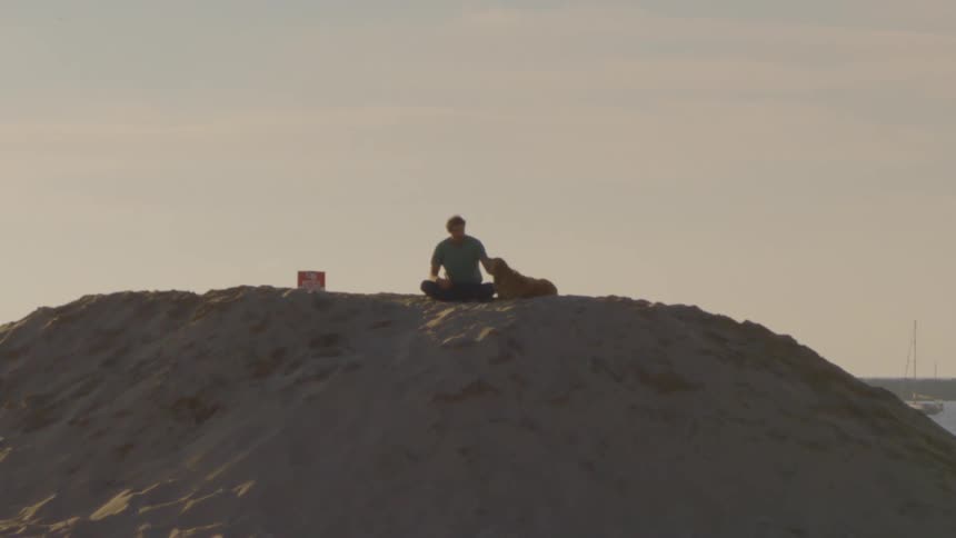 A man sits crosslegged atop a large sand dune at dusk stroking a brown dog sitting beside him while a sailboat floats on the water in the background under a hazy sky