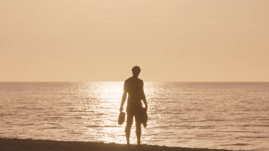A silhouette of a man stands on the beach at sunset holding his sandals while looking out at the ocean