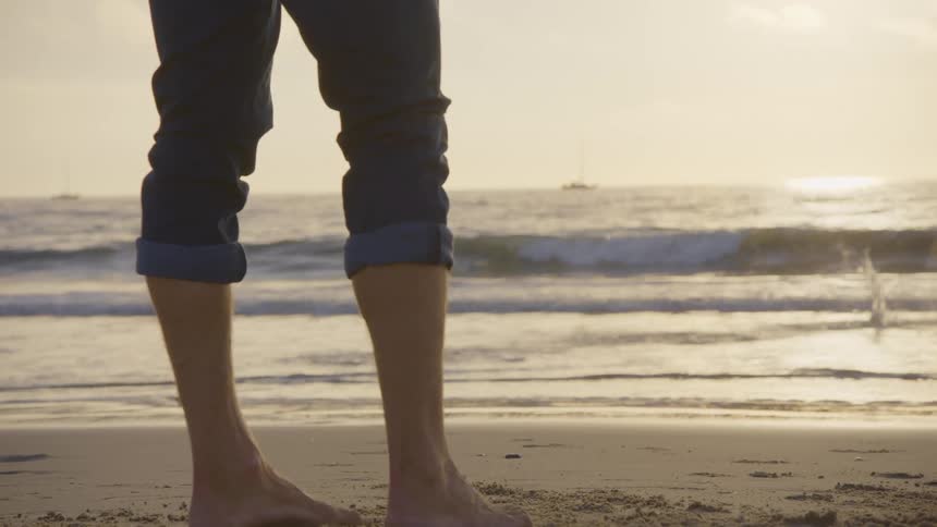 A persons legs are standing on a beach as a golden retriever runs on a beach into the ocean