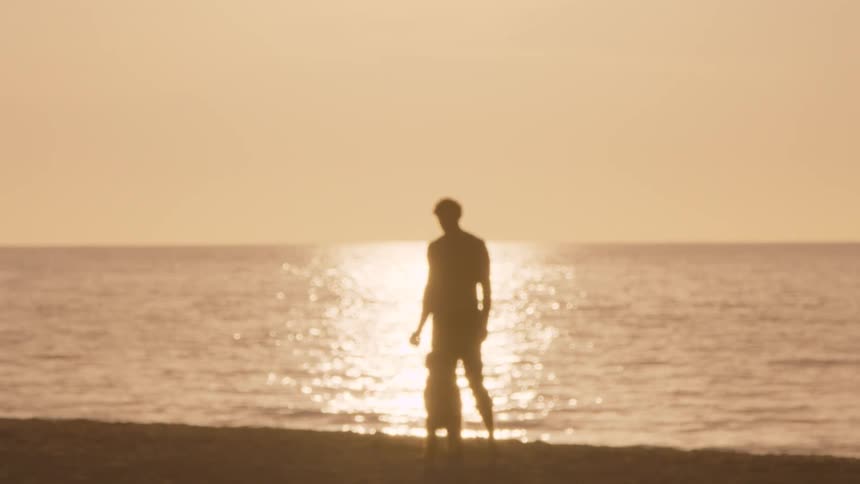 Man stands on the sand facing the sea with low sun reflecting on the water