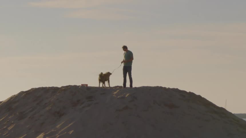 A man stands atop a large sand mound with a mediumsized dog on a leash the pair is silhouetted against a pale hazy sky as the dog moves around its owners feet