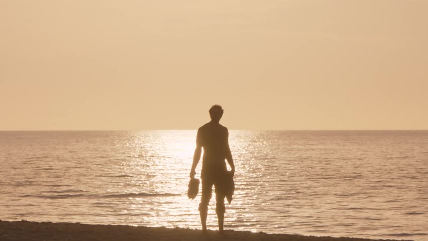 A man standing on the beach watching the setting sun above the ocean the orange sky glowing as sunlight reflects across the water while he holds his shoes in one hand