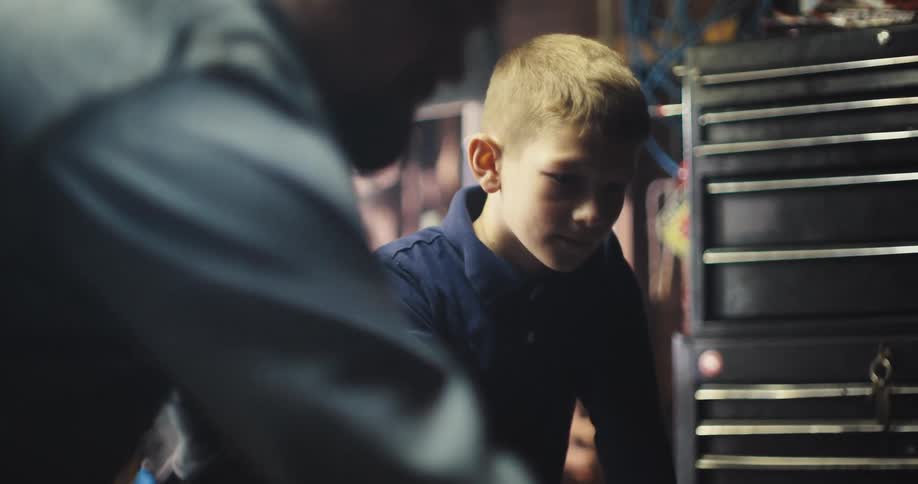 A young boy walking into a garage where his father is fixing the car the boy standing nearby watching him work