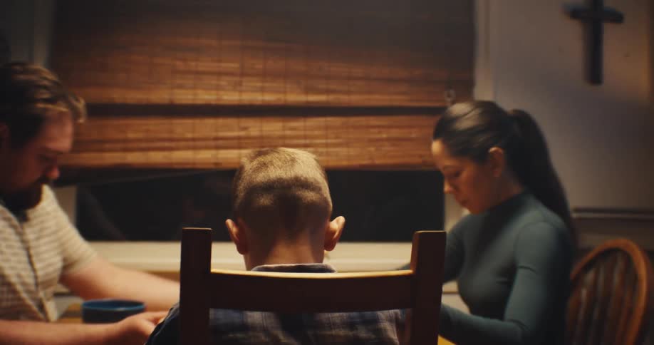 A family bows their heads in prayer at a small dinner table under a wall mounted crucifix