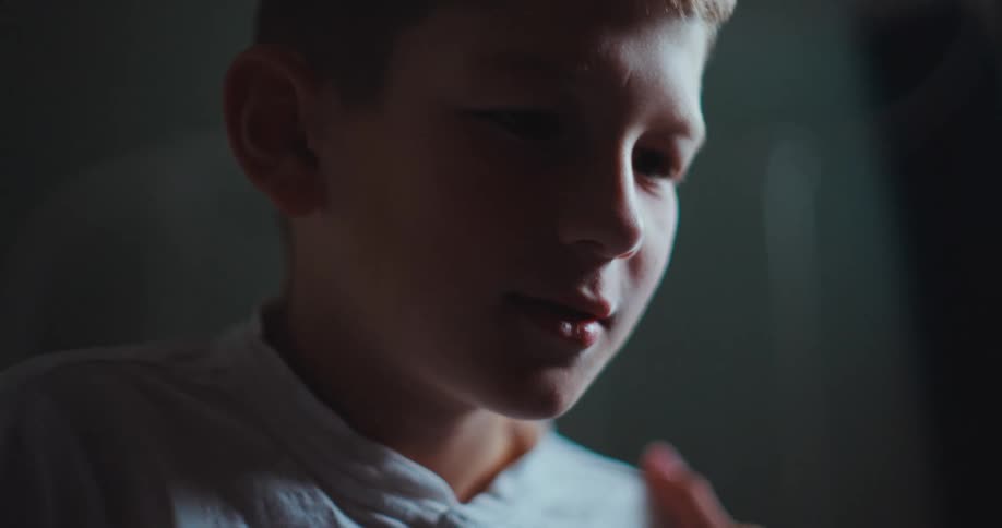 In a dimly lit room a young boy wearing a white shirt gazes downward with a focused and slightly somber expressionas he looks up and speaks someones hand reaches into the frame to gently brush the hair away from his forehead