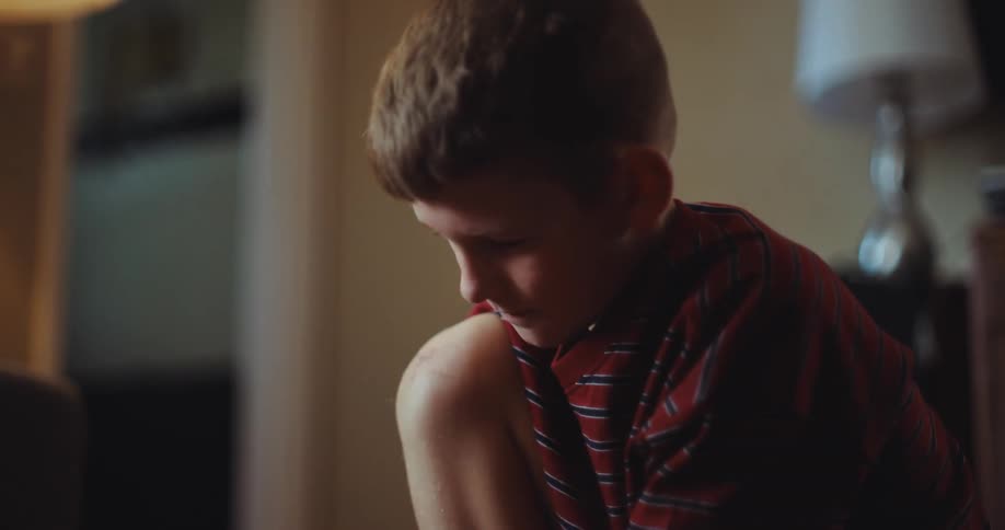 A young boy in a striped shirt sits on the floor deeply focused on playing with his dinosaur toys he gently moves a green dinosaur figure around while occasionally glancing down at a small scrape on his knee