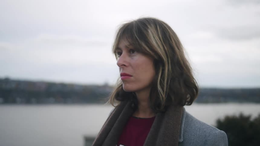 A woman with medium length hair and bangs wearing a grey coat and a brown scarf over a maroon shirt looks out over a body of water and a distant cityscape on an overcast day
