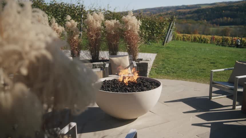 Outdoor fire pit burning on a patio with chairs and a rural landscape in the background