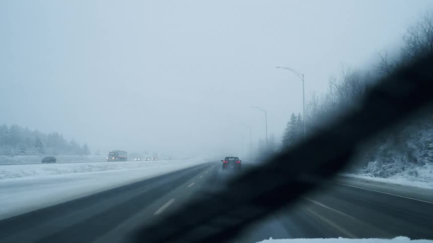 A car travels down a multilane highway surrounded by a landscape of snowcovered trees and ground during a heavy winter stormvisibility is limited by thick fog and falling snow as windshield wipers clear the frame and other vehicles drive cautiously a