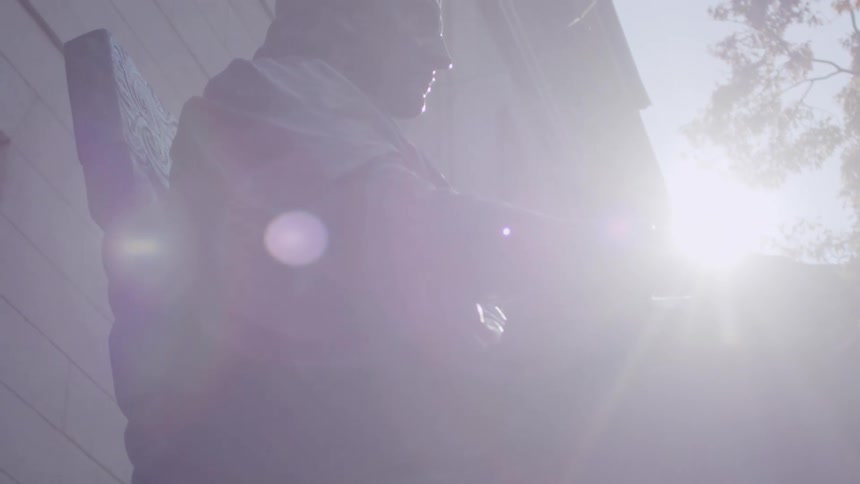 This video shows a lowangle shot of a dark bronze statue of a seated man holding a large book bright sunlight flares behind the statue partially obscuring the figure as the camera focuses on its profile against a building and a tree