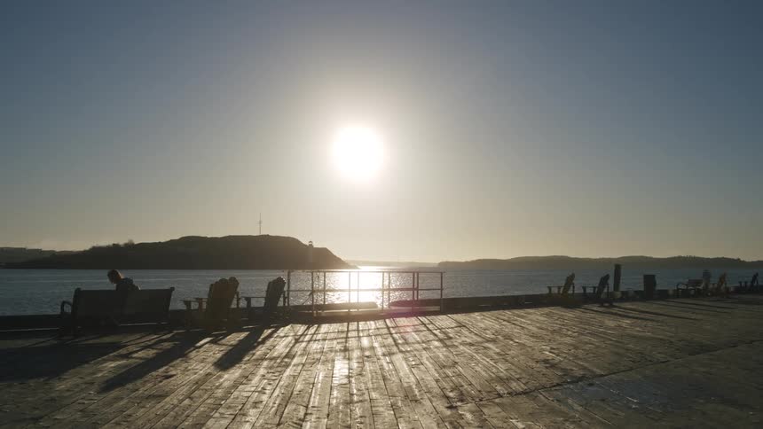 Wooden pier with empty chairs beside a body of water under the low sun on the horizon
