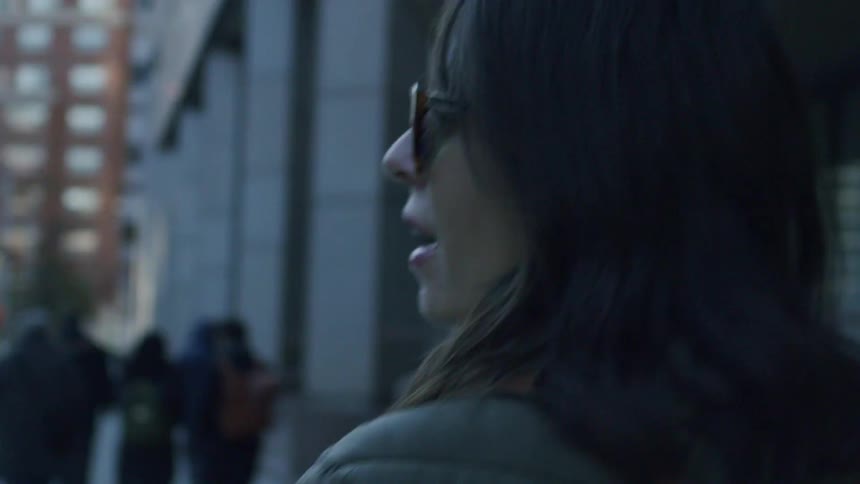 A woman with dark hair and a green puffer jacket walks through a city at dusk she briefly looks over her shoulder toward the camera before continuing her walk down the sidewalk