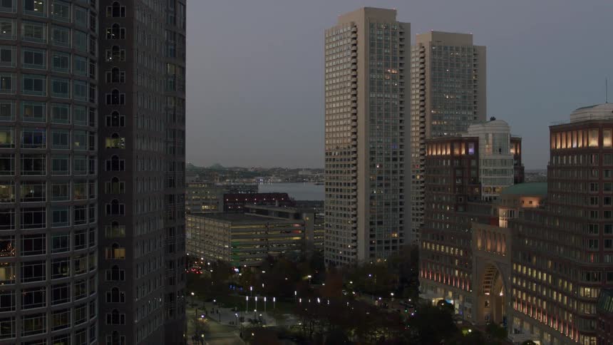 The footage shows a steady highangle view of a city skyline at dusk featuring several tall residential and commercial buildings illuminated by interior lights