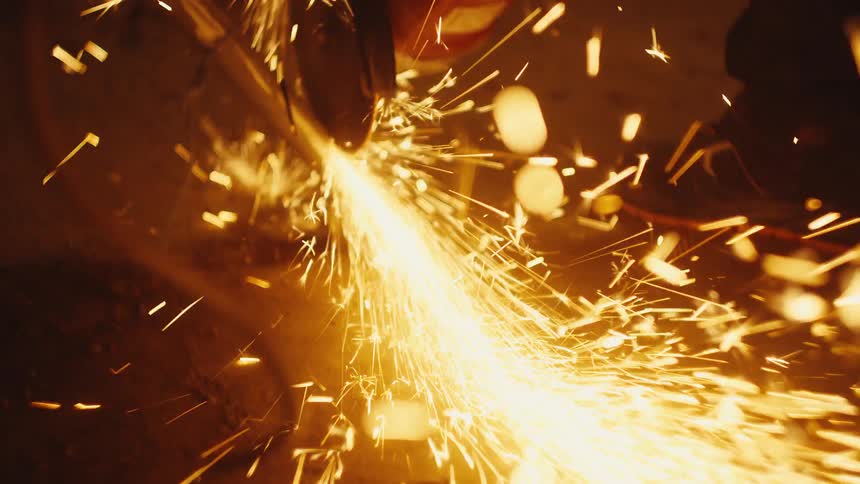 A power tool with a rotating abrasive disc is being used to cut through a metal pipe creating a heavy stream of bright orange sparks that illuminate the dark workspace