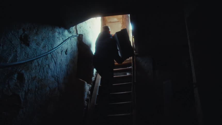 Person climbs stairs carrying a bulky object inside a dim interior lit from above