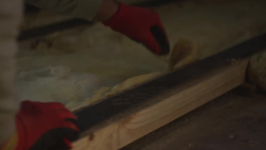 A worker wearing red and yellow gloves carefully presses thick yellowish mineral wool or fiberglass batts into the spaces between structural beamsthe scene focuses on the repetitive manual labor of fitting the insulation snugly against the timber to i