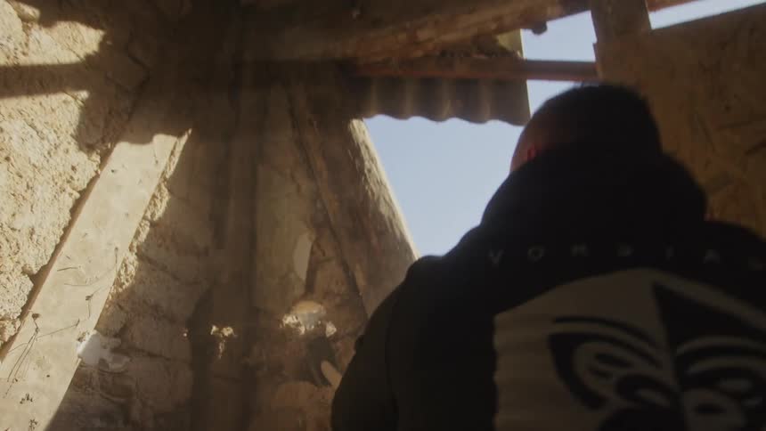 A man in a black hoodie repeatedly swings a sledgehammer to demolish a stone wall inside a dilapidated structuredust and debris fly into the air as the heavy tool strikes the weathered masonry under bright natural light