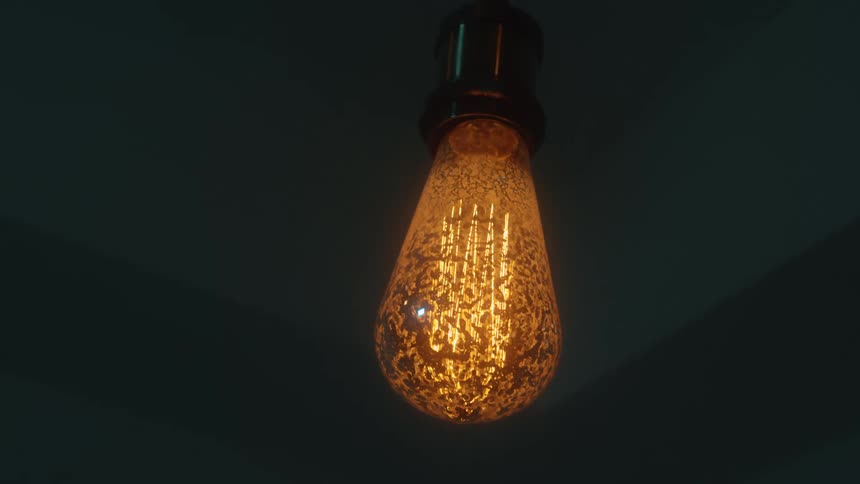 An orange edisonstyle light bulb hangs from a dark ceiling glowing with a warm and intricate filament patternthe light subtly flickers and dims against the moody shadowed background of the room