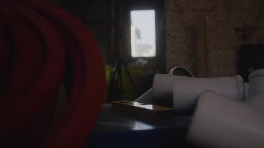 In a dimly lit workshop the view slowly orbits around a collection of white pvc pipe fittings and a yellow level resting on a dusty shelf
