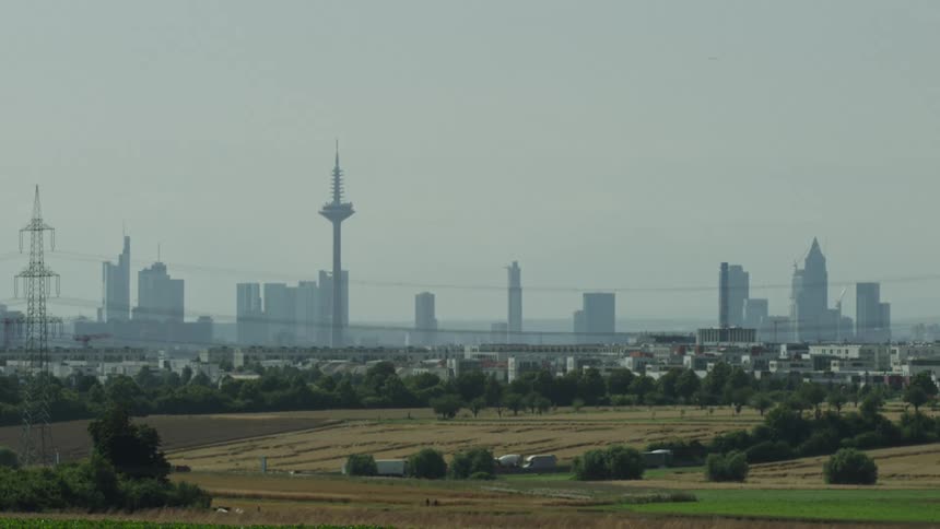 A wide landscape view shows a hazy city skyline in the distance featuring a prominent telecommunications tower and several skyscrapers in the foreground a rural setting consists of green trees utility poles and golden fields under a pale sky