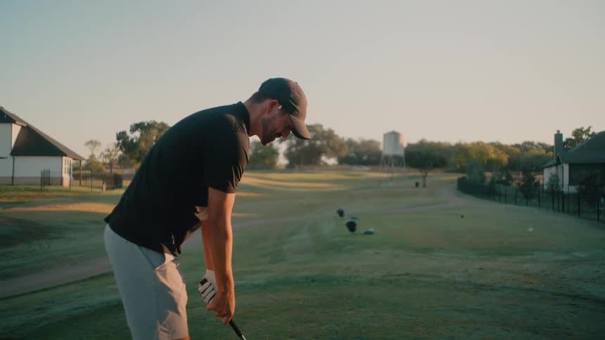A man wearing a black shirt and grey shorts practices his golf swing on a grassy tee box during the early morning although he swings his club multiple times he intentionally does not hit the golf ball resting on the ground in front of him