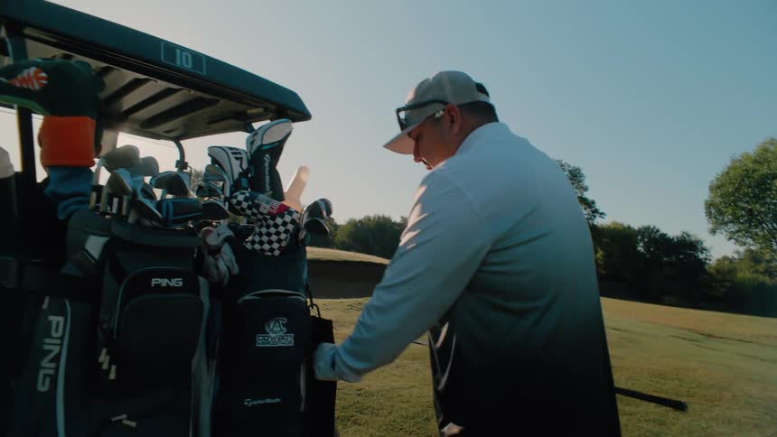 A man wearing a grey gradient shirt and a hat walks toward a golf cart parked on a grassy course and pulls a club out of his black golf bag under a bright clear sky