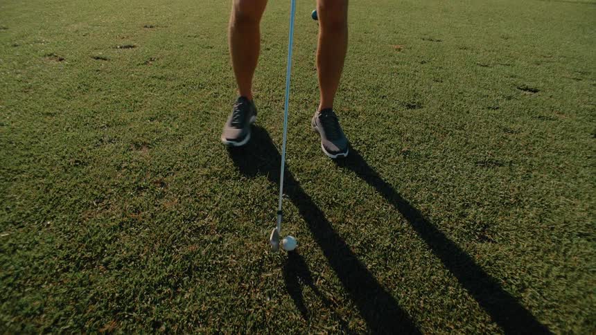 A golfer stands on a sunlit green carefully aligning the head of their club behind a white golf ball the low morning sun casts a long distinct shadow of the player across the grass as they prepare for the shot