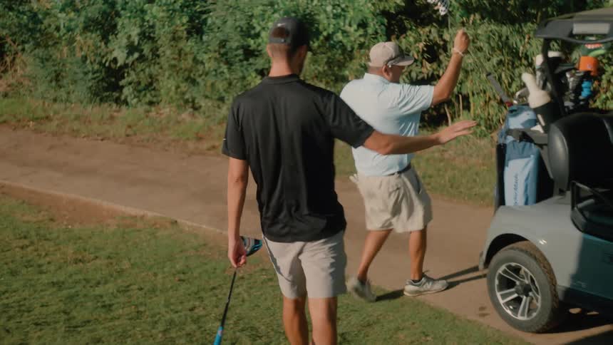 Two men carrying golf clubs walk to a golf cart and put them in their golf bags