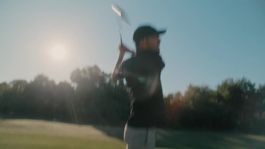A golfer addresses a ball on the grass and takes a swing sending the ball high into the airthe scene shifts to show the golfer completing his followthrough while watching the balls flight against a sunny treelined backdrop
