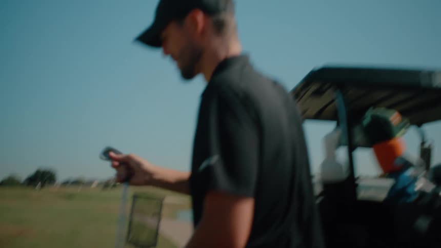 A male golfer takes a club from the golf cart and walks toward the golf ball under the sun