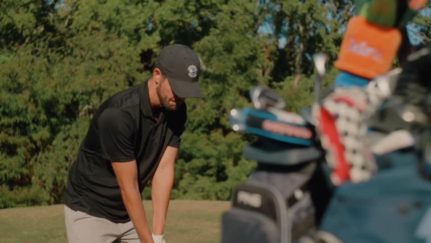 A male golf player swings his club at midday sending the ball flying across the course with a set of clubs resting in a nearby cart