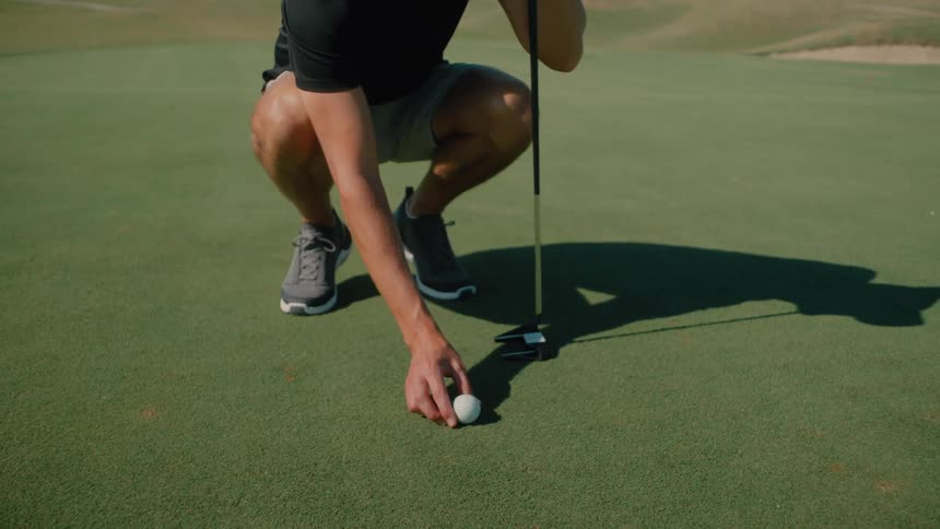 A man in a black shirt and grey hat crouches on a golf green while leaning on his putter he reaches down to place a golf ball on the grass before standing up to prepare for his next shot