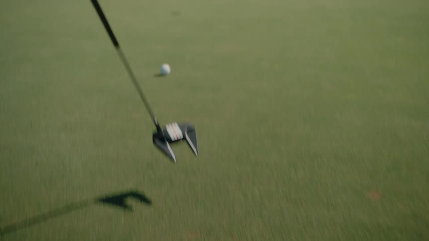 A golfer uses a malletstyle putter to hit a white golf ball across a manicured green toward a hole while houses and trees are visible in the blurred background under bright daylight