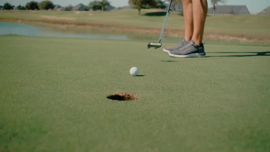 A golfer putts a white ball across a green watching it roll directly into the holeafter the successful shot the golfer walks forward and reaches down into the cup to retrieve the ball