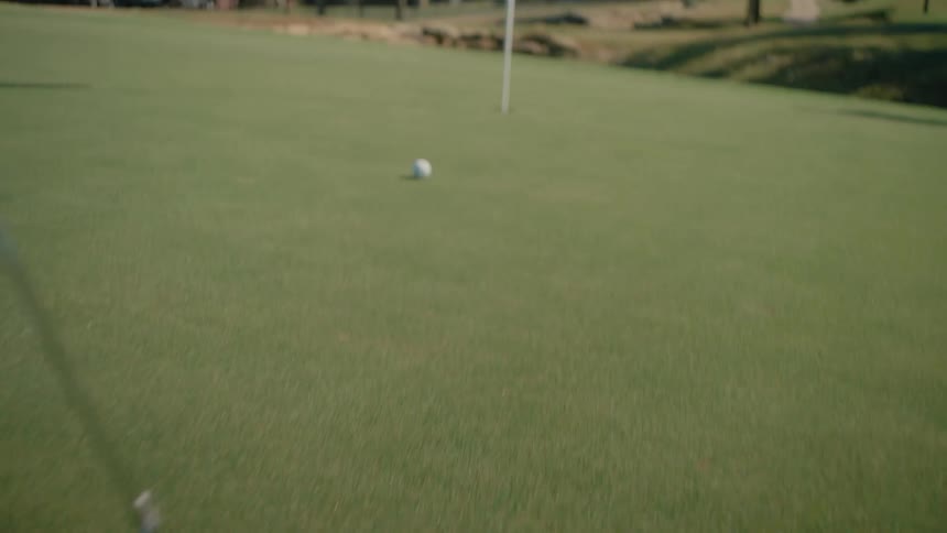 A golfer uses a silver putter to strike a white golf ball across a manicured green the ball rolls steadily toward the hole as the camera follows its path across the grass