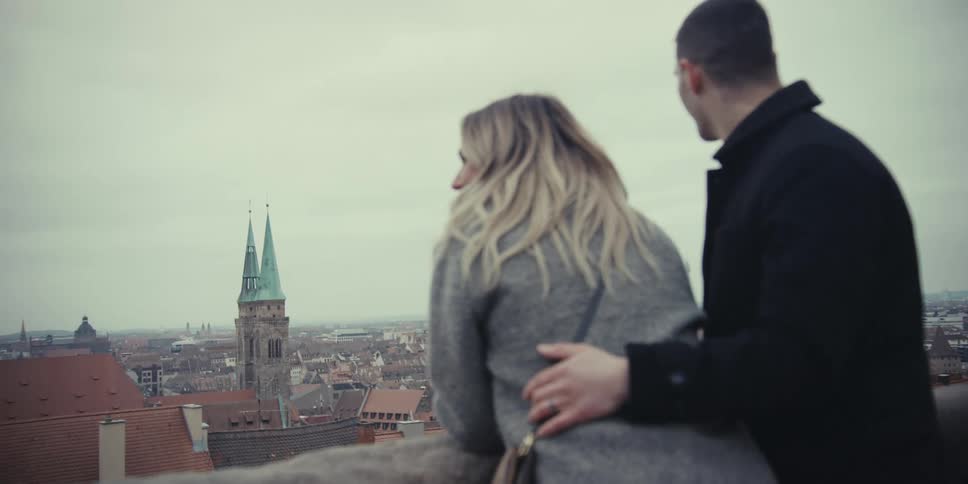 Man and woman standing together with his arm around her and he kisses her forehead as she leans against the railing overlooking neighborhood buildings below