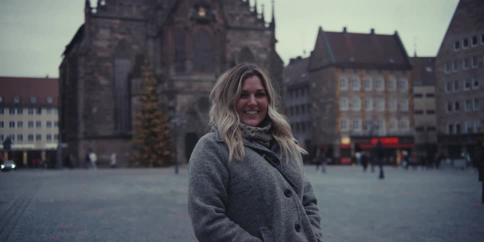 A blonde woman in a grey coat stands in a historic city square her warm smile slowly settling into a gentle confident gaze at the camera