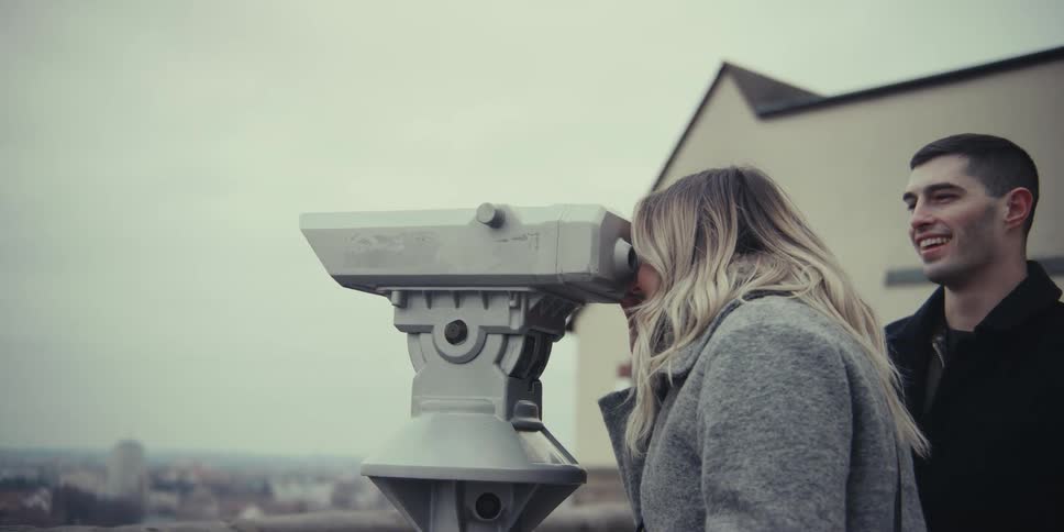 A young man and woman stand at a city overlook and the man looks through a stationary telescope and then turns to smile at his companion before she takes a turn peering through the lenses and they both laugh while enjoying the view together