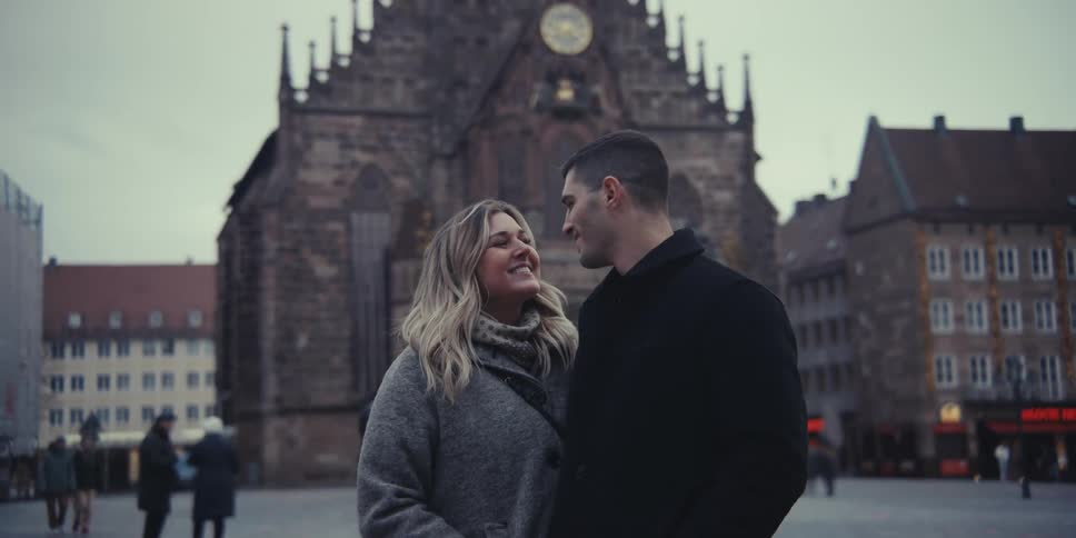 A man and a woman stand in a city square in front of a large gothic church they look at the camera turn to smile at each other share a brief kiss and then return to posing together as people walk by in the blurred background