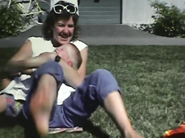 In a grainy home video a mother playfully tickles and kisses her young son on a sunny lawn and they both burst into joyful laughter before she waves