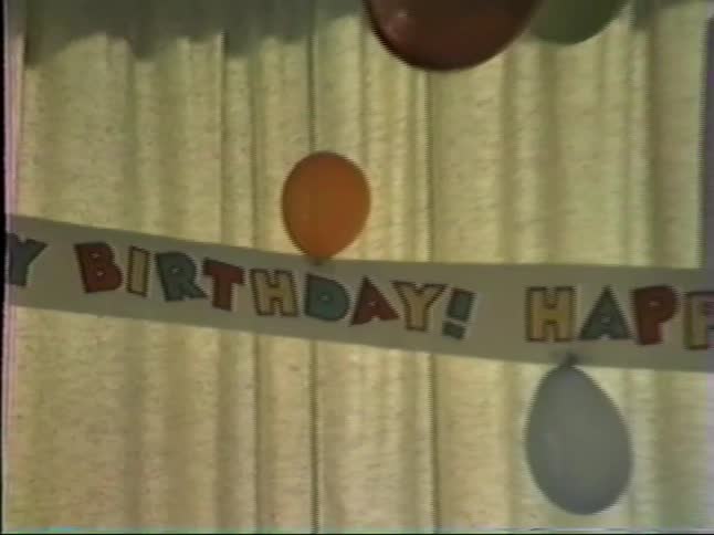 A grainy home video pans slowly across a colorful paper happy birthday banner and balloons strung up in front of a window ready for a celebration