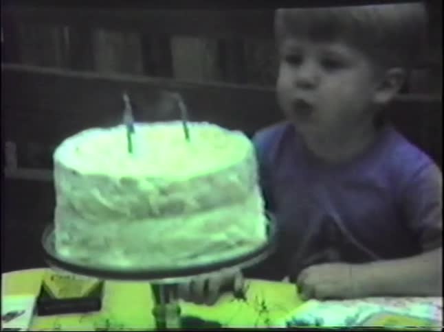 From a grainy home video a young boy with blonde hair takes a big breath and successfully blows out the three candles on his birthday cake