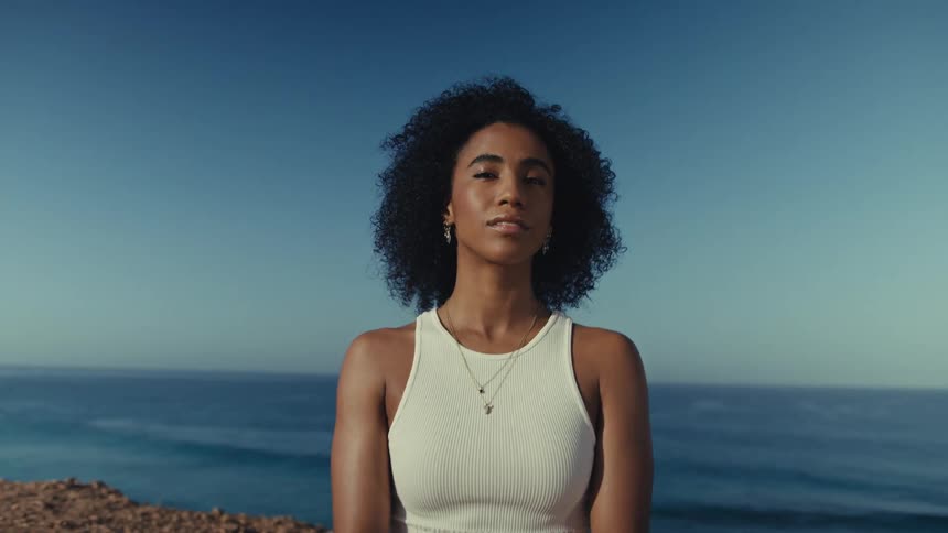 Young adult black woman sitting on a yoga position next to a cliff next to the ocean outdoors during day time