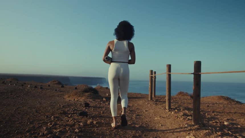A woman with dark curly hair walks away from the camera along a dirt path overlooking the ocean she wears white athletic gear and carries a rolledup yoga mat as she heads toward a scenic cliffside