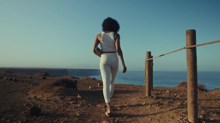 Woman walking along a coastal path holding a training mat with the ocean in the background