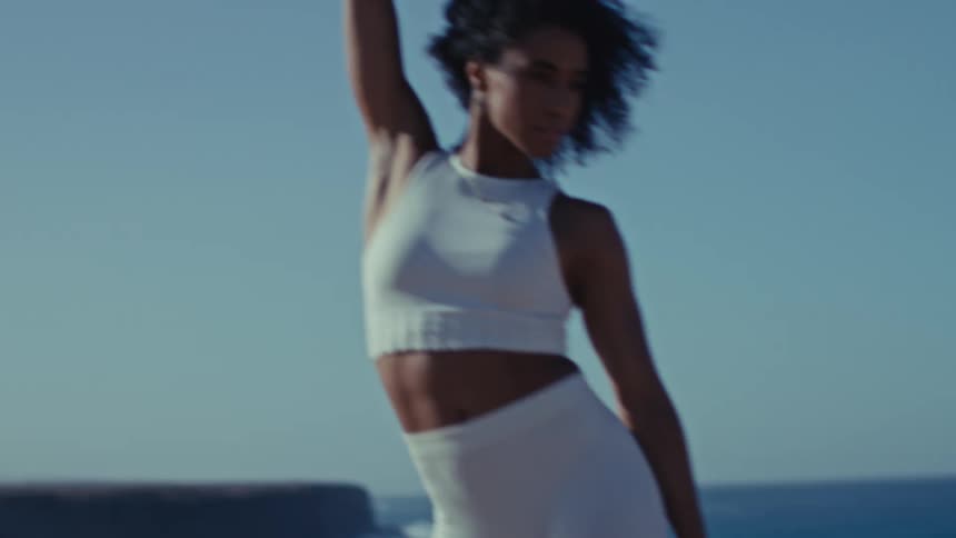 A young woman dances on a hill along the shoreline at sunset her long curly hair moving with each motion the white top and white yoga pants catching the warm glow