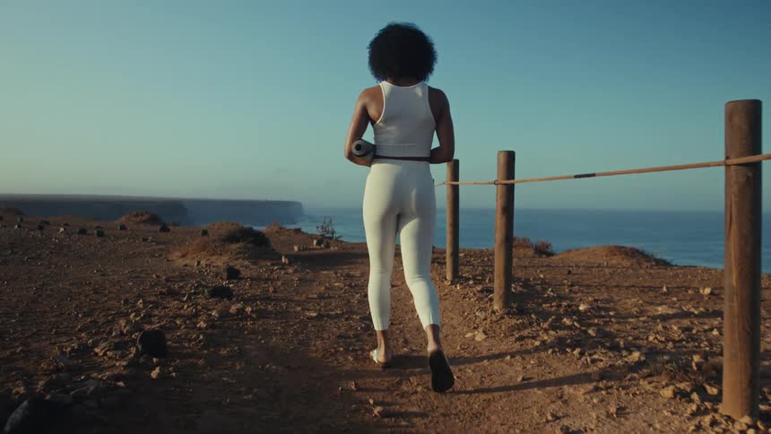From behind a woman in athletic wear confidently walks along a dusty cliffside trail carrying a yoga mat as she soaks in the breathtaking ocean view