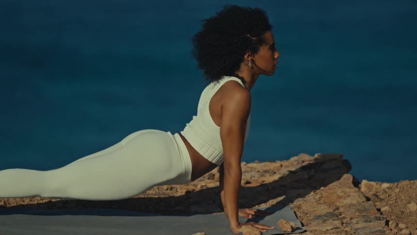 Woman holds a yoga pose on a mat atop a cliff with the ocean in the background