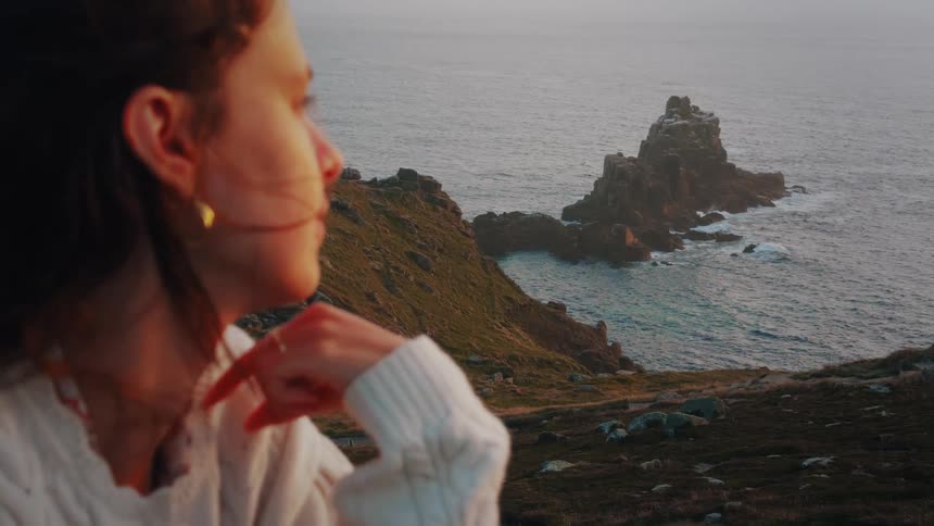 A woman with dark hair sits on a grassy cliffside resting her chin on her hand as she looks out toward the oceanin the distance a large jagged rock formation rises from the hazy water under the soft light of a setting sun