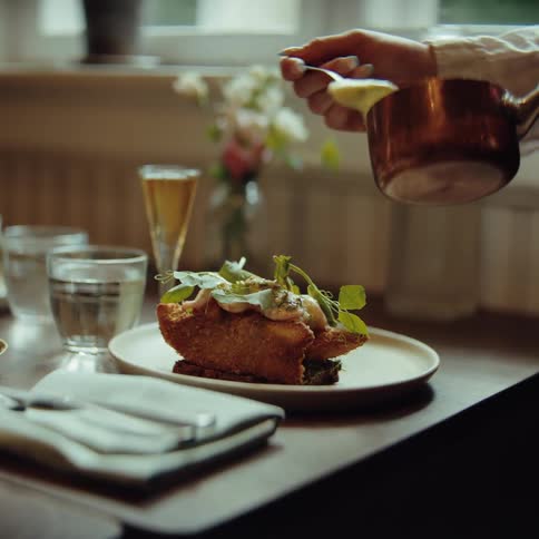 A chef meticulously drizzles a creamy sauce from a small copper pot onto a plated dish the meal consists of a goldenbrown breaded protein garnished with fresh greens in an elegant dining setting